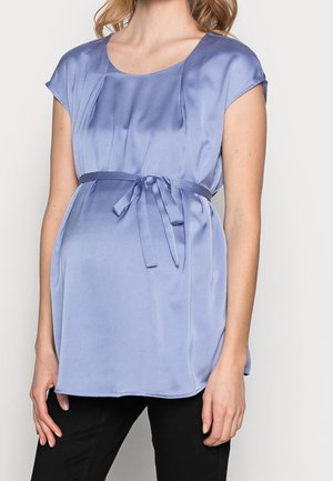 Mujer vistiendo una blusa de maternidad sin mangas de satén azul claro con un lazo atado sobre la barriga de embarazo, combinada con pantalones negros.