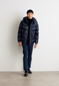 Navy pufferjacka med huva, dragkedjefickor och en glänsande textur. I kombination med svart polotröja, mörka jeans och svarta skor.