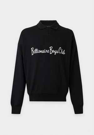 Man draagt een zwarte polo met lange mouwen met de tekst "Billionaire Boys Club", donkere losse spijkerbroek met een luipaardpatch en witte sneakers.