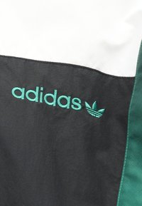 Melnbalta adidas jakna ar zaļu izšūtu logotipu un akcentiem, gluda tekstūra, ar sportisku krāsu bloku dizainu un kontrastējošu apdari.