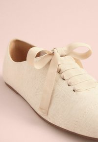 Chaussure en toile beige avec un bout rond, surface texturée et fermeture à lacets plate. Présente un nœud en tissu assorti à l'avant.