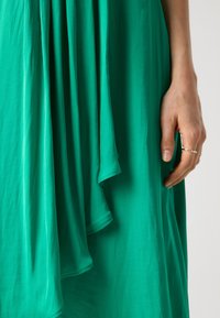 Green, flowing dress with layered fabric featuring smooth texture. Hand with a silver ring holds the hem, displaying gentle draping.