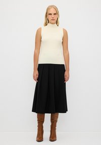 Lichte crème mouwloze top met opstaande kraag, gecombineerd met een lange zwarte geplisseerde rok en bruine kniehoge laarzen. Soepele texturen, elegante silhouet.