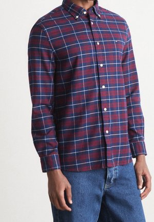 Homme portant une chemise à manches longues à carreaux rouges, bleu marine et blancs, ainsi qu'un jean bleu, debout les bras détendus.