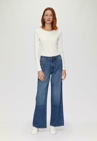 Weißes Langarmoberteil, kombiniert mit weit geschnittenen blauen Jeans. Die Jeans sind hochgeschnitten und haben am Saum eine leichte Fransenoptik, getragen mit weißen Sneakers.