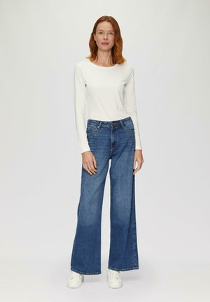 Weißes Langarmoberteil, kombiniert mit weit geschnittenen blauen Jeans. Die Jeans sind hochgeschnitten und haben am Saum eine leichte Fransenoptik, getragen mit weißen Sneakers.