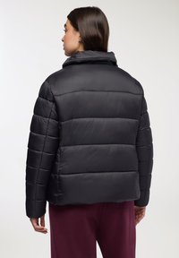 Kobieta stojąca tyłem, ubrana w czarną pikowaną kurtkę puchową z wysokim kołnierzem i bordowe spodnie, na jasnym tle.