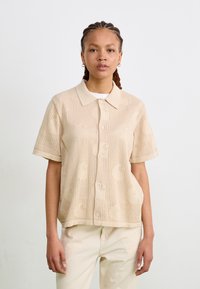 Beige knitted polo shirt with short sleeves, featuring a subtle textured pattern and front buttons. Smooth fabric with a relaxed fit.