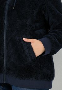 Hånd i sidelommen på en navyblå fuzzy zip-up jakke med ribbede manchetter og talje, båret over et par blå jeans.