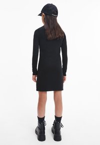 Abito nero a maniche lunghe con una vestibilità aderente, abbinato a un berretto nero e stivali neri robusti. Tessuto liscio, lunghezza fino a metà coscia.