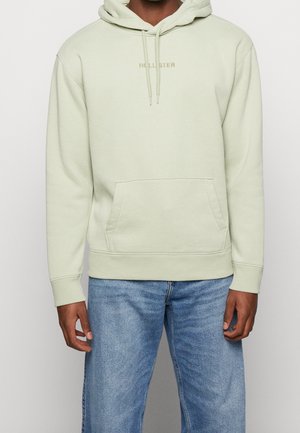 Personne portant un sweat à capuche Hollister beige clair avec une poche avant et des cordons, assorti à un jean bleu clair sur un fond uni.