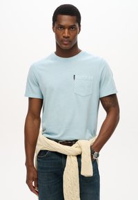 Camiseta de algodón azul claro con un bolsillo en el pecho. El modelo lleva un suéter de punto trenzado crema atado a la cintura y jeans oscuros con un cinturón marrón.