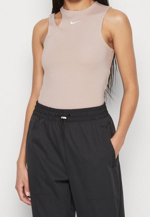 Nainen pukeutuneena hihattomaan, olkapään läheisyydessä pieneen aukkoon varustettuun beigeen Nike-toppiin ja mustiin, taskuissa kädet oleviin vetoköysihousuihin.