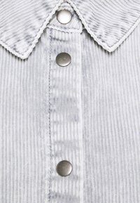 Gros plan d'une chemise en velours côtelé blanc à rayures verticales, trois boutons-pression métalliques et un col aux bords cousus.