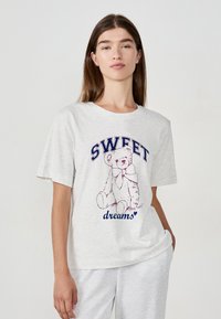 Vrouw in een lichtgrijze t-shirt met een teddybeer grafiek en de tekst "SWEET dreams" staande tegen een effen lichte achtergrond.