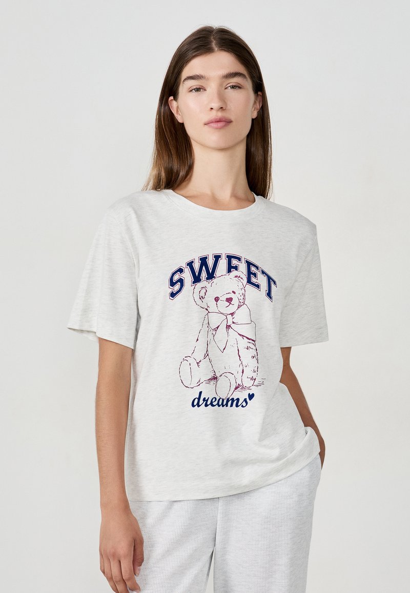Vrouw in een lichtgrijze t-shirt met een teddybeer grafiek en de tekst "SWEET dreams" staande tegen een effen lichte achtergrond.