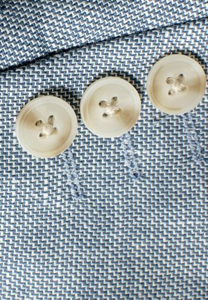 Tres botones crema con un detalle en forma de flor están cosidos sobre una tela tejida azul y blanca con textura, que presenta un patrón diagonal.