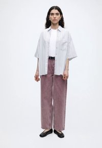 Jeune femme portant une chemise ample à manches courtes rayée par-dessus un t-shirt blanc, un jean rose poussiére à jambes larges, des ballerines noires, debout devant un fond uni.