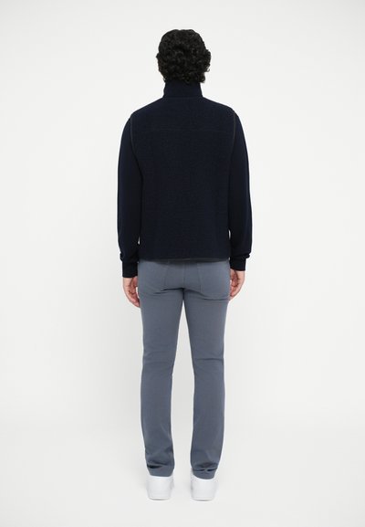 Marineblaue Fleece-Pullover mit hohem Kragen, kombiniert mit grauen Slim-Fit-Hosen und weißen Sneakers, von hinten gesehen. Glatte Textur, minimale Details.