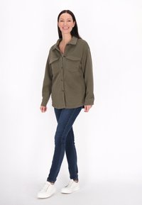 Giacca camicia trapuntata olivo con due tasche frontali, chiusura a bottoni, abbinata a jeans skinny scuri e sneaker bianche. Modella in piedi su uno sfondo bianco.