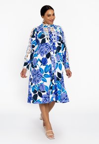 Blauwe bloemen midi-jurk met kantaccenten, knoopsluiting aan de voorkant, lange mouwen en een relaxte pasvorm. Heeft een witte achtergrond met donkere bladeren.
