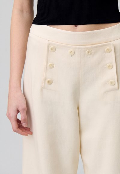 Pantalons larges crème en tissu texturé, avec deux poches latérales et six boutons décoratifs sur le devant.