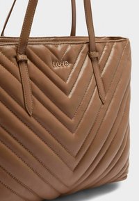 Borsa a mano in pelle marrone trapuntata con motivo a zigzag, dotata di due manici e un dettaglio con logo dorato sulla parte anteriore. Texture liscia, forma strutturata.