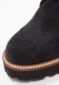 Gros plan sur l'avant d'une chaussure en daim noir avec perforations brogue, surpiqûres brunes sur la trépointe et semelle épaisse en caoutchouc noir.