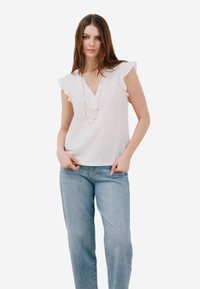 Camicia bianca con maniche a volant, scollatura a V e dettagli di bottoni, indossata con jeans azzurri. Modella accessoriata con una collana colorata.
