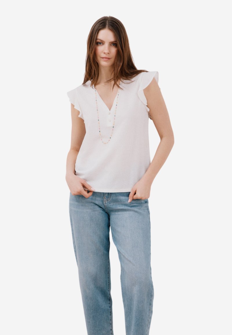 Camicia bianca con maniche a volant, scollatura a V e dettagli di bottoni, indossata con jeans azzurri. Modella accessoriata con una collana colorata.