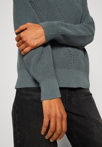 Sweter dzianinowy w kolorze morskim, z teksturowanym wzorem, prążkowanymi mankietami i dołem, o luźnym kroju. Uzupełniony ciemnymi spodniami.