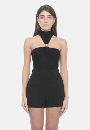 Donna che indossa una tuta nera senza maniche con collo all'americana e dettaglio ad anello dorato sulla scollatura, in piedi davanti a uno sfondo semplice.