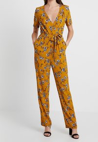 Žlutý květinový overal s krátkými rukávy, překládaným V výstřihem, stáhnutým pasem, dvěma bočními kapsami a rovnými nohavicemi. Obsahuje modré a bílé květy.