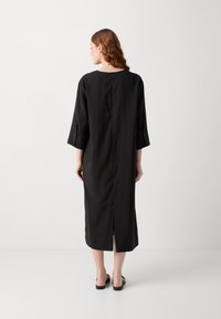 Robe noire à manches longues, coupe droite, fente au dos et fermeture par bouton au cou. Fabrication en tissu lisse. Design minimaliste.
