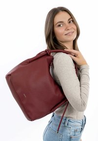 Raudona odinė kuprinė su tekstūruotu paviršiumi, minkšta forma, raišteliu uždaroma ir minimaliais prekės ženklais, nešiojama ant vieno peties.