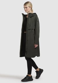 Olive green waterproof jacket with a hood, front snap closures, and side pockets. Paired with black leggings and black sneakers.