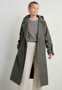 Olijfgroen trenchcoat met dubbele knopen, gezette revers en lange mouwen, gecombineerd met een gestreepte trui en lichtgekleurde broeken.