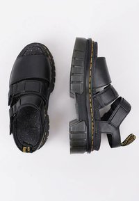 Des sandales noires avec des sangles en Velcro, des semelles en caoutchouc épaisses et des accents texturés. La semelle présente un détail de couture jaune.