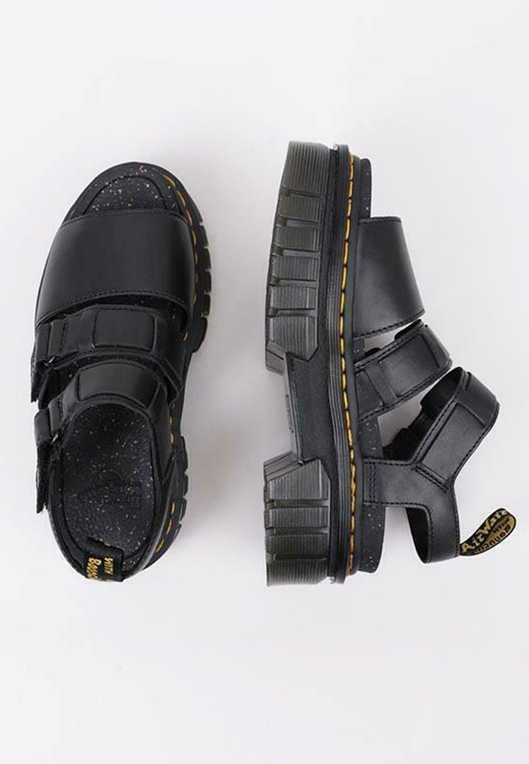 Sandalias negras con correas de Velcro, suelas de goma gruesa y acentos texturizados. La suela presenta un detalle de costura amarilla.