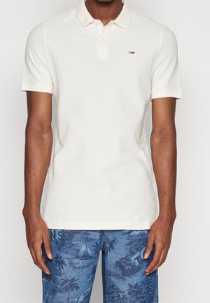 Homme portant un polo blanc à manches courtes avec un petit logo sur la poitrine gauche et un pantalon en jean bleu imprimé.