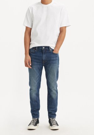 Homme portant un t-shirt blanc uni à manches courtes, un jean bleu et des baskets montantes noires, debout avec une main dans la poche.