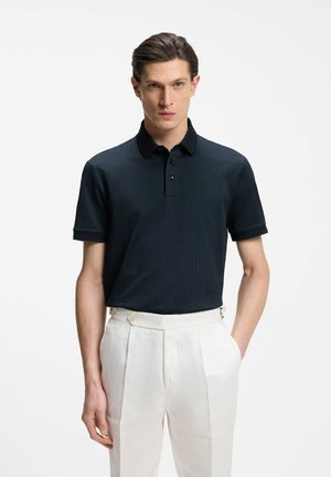Man wearing black short-sleeve polo shirt and high-waisted white pleated trousers, standing with one hand in pocket against plain background.