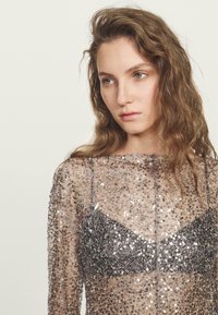Top lungo a maniche lunghe trasparente adornato con paillettes argentate, con un reggiseno argentato texturizzato sotto. Sfondo neutro.
