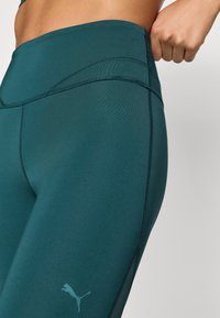 Teal színű, magas derekú leggings, sima textúrával, kontrasztos varrásokkal és kis logóval az alsó lábszáron. Nyújtható anyag, amely alkalmas edzésekhez.