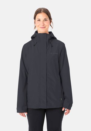 Donna che indossa una giacca impermeabile nera con cappuccio, cerniera frontale e polsini regolabili, in piedi contro uno sfondo chiaro e uniforme.