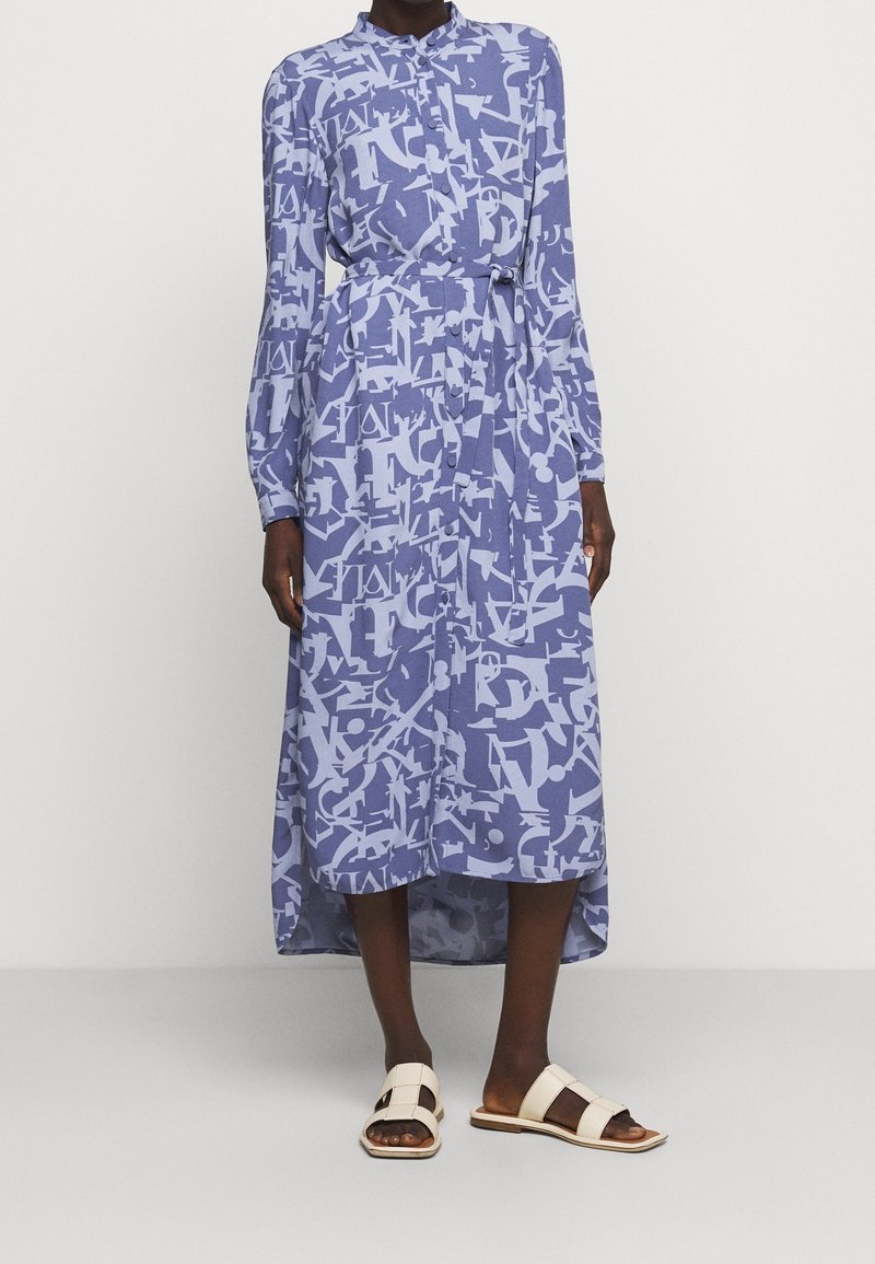 Lichtblauwe jurk met lange mouwen en een witte grafische print. Heeft een kraag, een tailleriem en een langere achterzoom. Paar crèmekleurige sandalen.