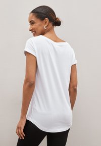 Femme avec les cheveux en chignon portant une chemise blanche à manches courtes et un pantalon noir, souriante et légèrement tournée sur le côté devant un mur uni.