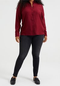 Röd skjorta med knappar och en subtil mönstring, tillsammans med svarta slim-fit jeans och svarta loafers. Texturen ser slät ut, med långa ärmar.