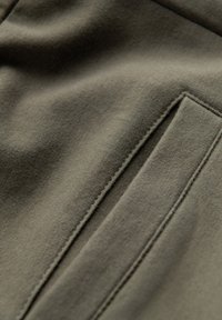 Olivgrüner Stoff mit einer glatten Oberfläche, einem Paspeltaschen und sichtbaren Nähten. Das Material ist dehnbar und figurbetont.