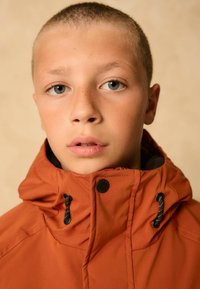 Pomarańczowa kurtka z kapturem i wysokim kołnierzem, z czarnymi akcentami i regulowanymi trokami. Prosty design o gładkiej teksturze.
